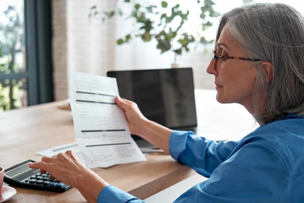 A silver-haired woman looking over financial documents at her desk, pondering her retirement plans.