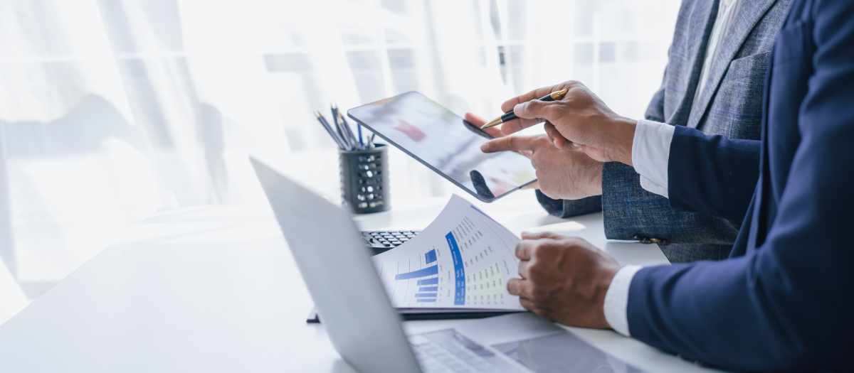 Two people in business attire look over data charts and laptop screens representing assets and wealth.
