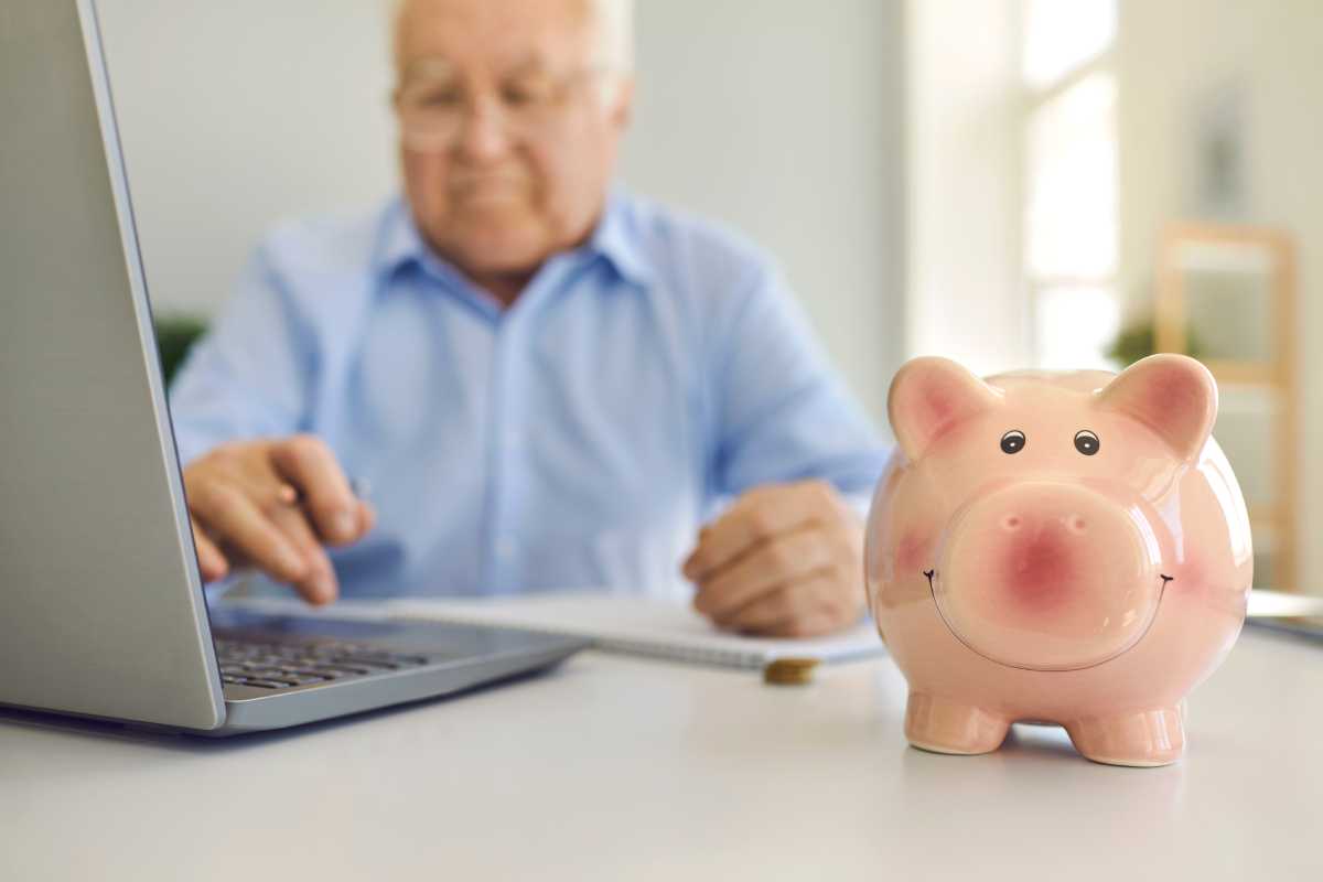 A senior looking at his laptop next to a piggy bank.