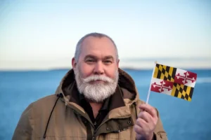 A man of retirement age holds a small Maryland state flag.