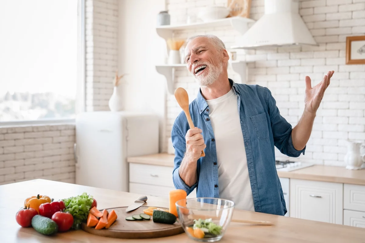 Cheerful older man smiles while holding a wooden spoon and cooking because his retirement planning checklist is all set
